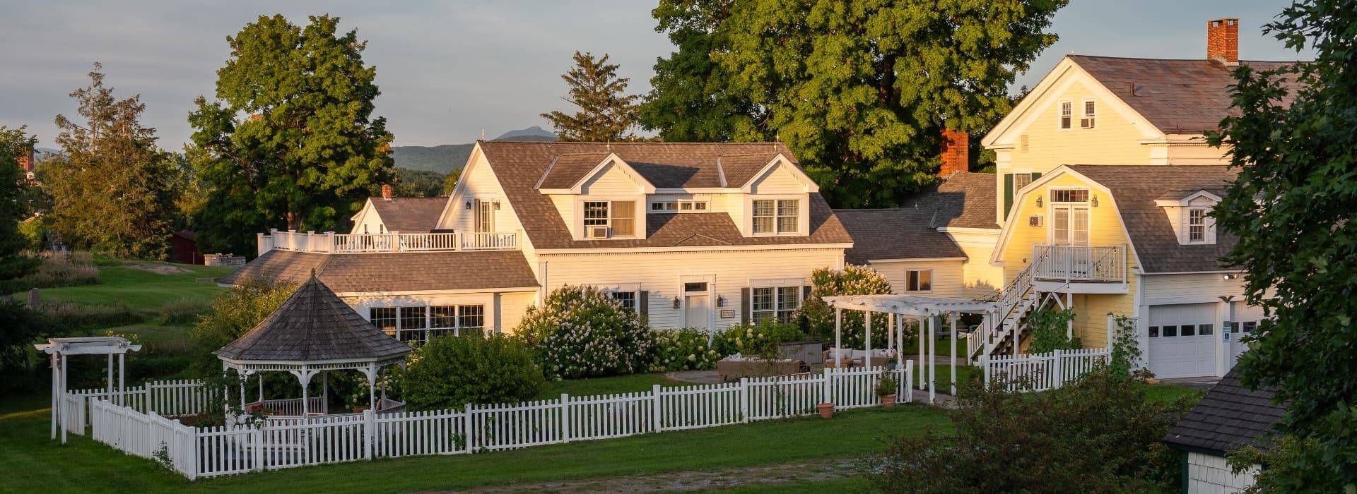 Exterior view of the property painted yellow with white trim and fence surrounded by green grass and trees