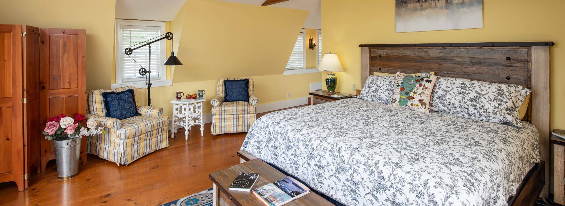 Bedroom with hardwood flooring, yellow walls, white trim, wooden headboard, and sitting area with two upholstered chairs